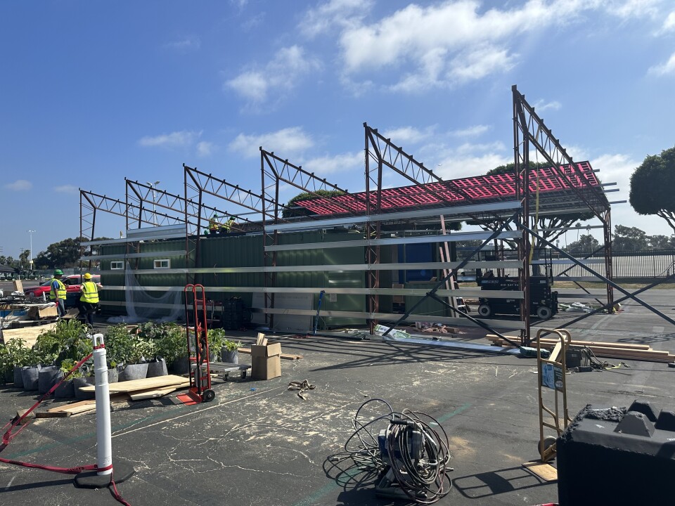 An in progress construction of the University of California Santa Cruz’s EcoHus sustainable home. The asphalt is scattered with various pieces waiting to be assembled, two workers are seen in the far left horizon line. In front of them are various potted plants lined up surrounding by plywood and a red dolly. To the right of them is the EcoHus, with six scaffolding like columns jutting out of the top of a green refurbished cargo container.