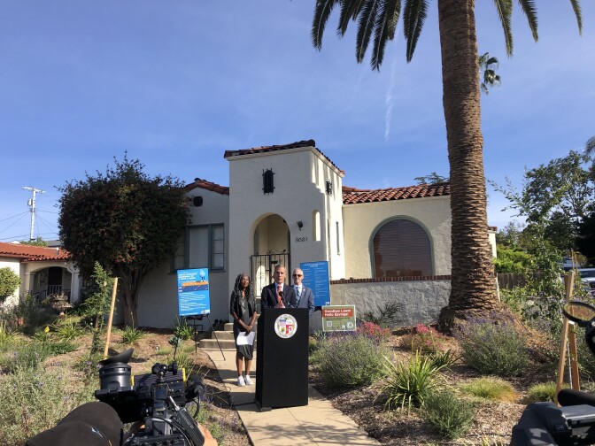 A wide shot of a man speaking at a black podium in front of an adobe-style home. A large palm tree is in the background and native plants are in the front yard. 