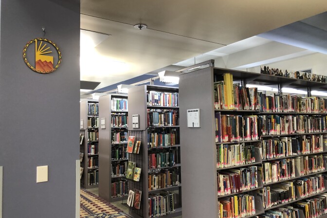 Three rows of grey bookshelves filled with different books in different colors at the Los Angeles Public Library. On the side of each shelf is a small white rectangle that serves as a sign saying what section the shelf is.
