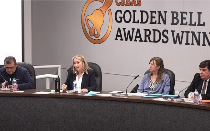 Four people are seated at a dais in front of a sign on a wall that reads Golden Bell Awards Winners. 