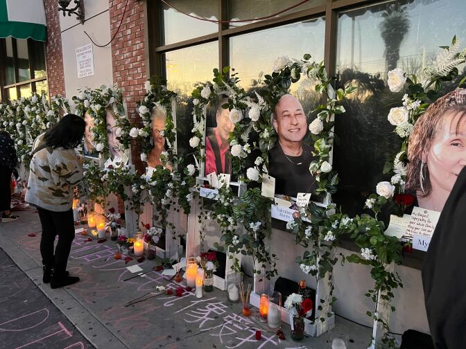 Large photos rest on easels rimming in white flowers with candles at the base. One women is bent over reading the messages.