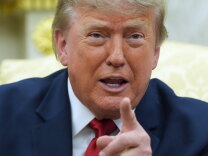 A man wearing a blue suit jacket and red tie is seated, pointing his left index finger while speaking.