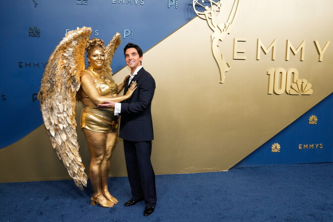 Meg Stalter is painted entirely gold and is dressed as the Emmys award while Paul W. Downs wears a dark suit and tie at the Emmy Awards red carpet.