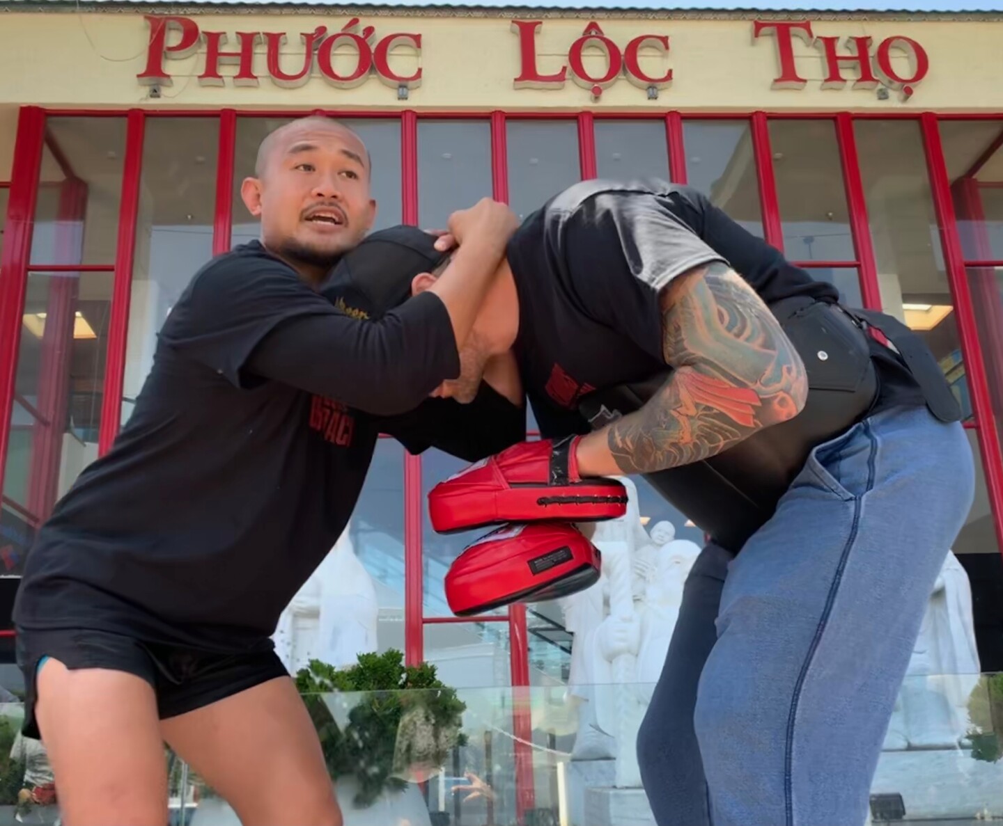 One man grips another man's head in front of a storefront reading Phuoc Loc Tho.