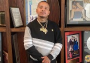 A man with dark tone skin has closely cropped hair. He has a thin mustache, wears several gold chains and has tattoos visible on his neck. He stands in front of shelves with records and recording artist photos.