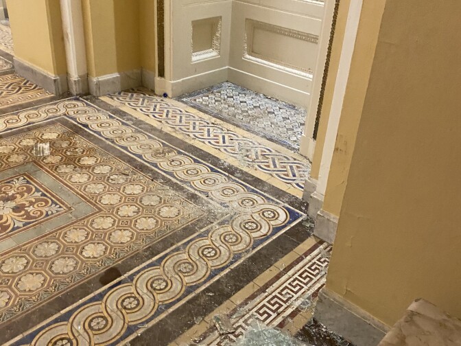 An interior room with tan walls and intricate tan, brown, and cream colored tiles. A window is broken on the right, with shattered glass and splintered wood on the floor around it.