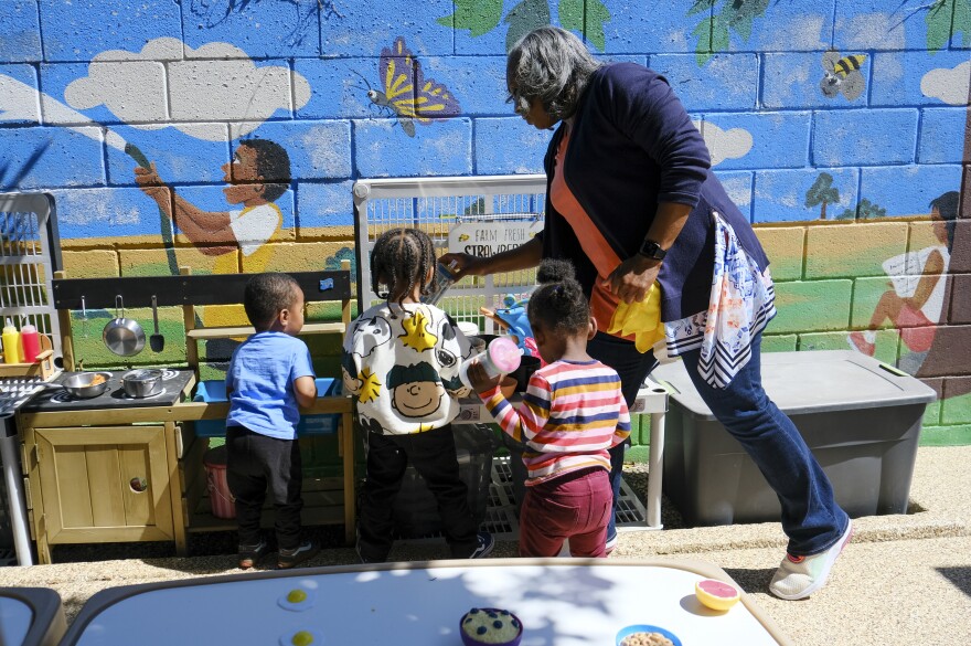 A Black woman in a pink shirt and cardigan leans over three children who are all waist-height or shorter playing at a toy kitchen. There is a mural of a blue sky, yellow butterfly and boy with dark skin tone and a garden hose.