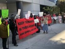 Tenants and organizers from Hillside Villa -- a Chinatown low-income housing complex -- stage a protest outside Los Angeles Housing Department on March 7, 2023 against general manager Ann Sewill. Two protesters in the center hold a red banner that says "KEEP TENANTS IN THEIR HOMES. ANN SEWILL MUST GO!" A young Asian woman speaks into a microphone on their right. Other protesters holding posters surround them.