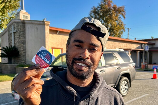A man with medium-dark skin tone wearing a black The North Face sweater and gray cap worn backwards holds up an "I Voted" sticker. 