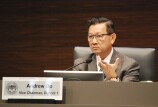 A man in a chair wearing a suit jacket, tie and glasses looks forward with a microphone in front of him. A sign in front has the official seal of the County of Orange and states "Andrew Do, Vice Chairman, District 1."