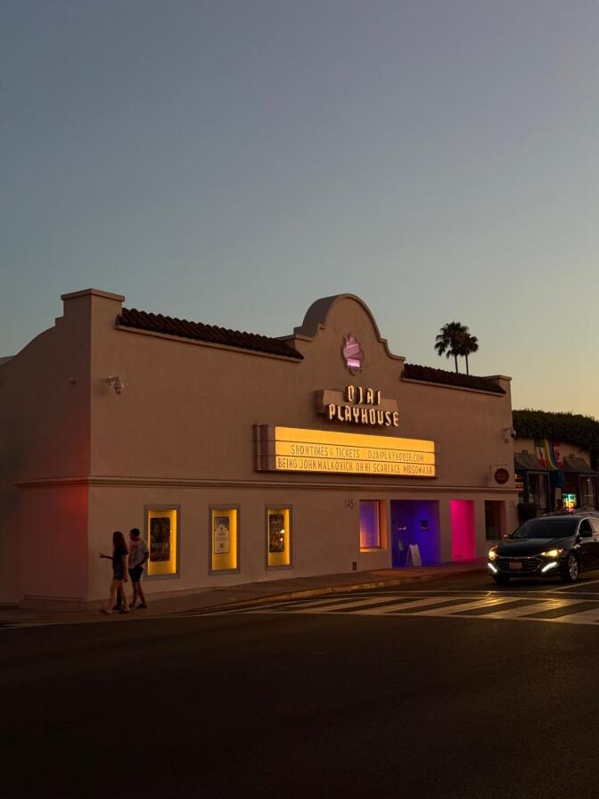 an adobe building with a sign reading "ojai playhouse" at dusk with two palm trees in the background, two people walking in front, and a car stopped in front of the building