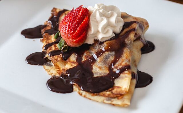 An image of a crepe sits on a white plate. The crepe is a light brown color and is covered with a dark brown chocolate sauce, a piped dollop of whipped cream and half a strawberry