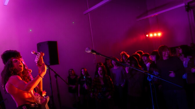 The band Saints of Sinners plays on stage under the glow of orange lights. The guitarist has long hair and is shirtless. 