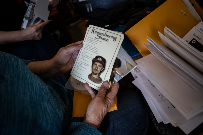 A pair of hands holds a memorial card that says Remembering Shane, which includes illegible text and drawing of a man with a mustache and goatee, wearing a baseball cap, 