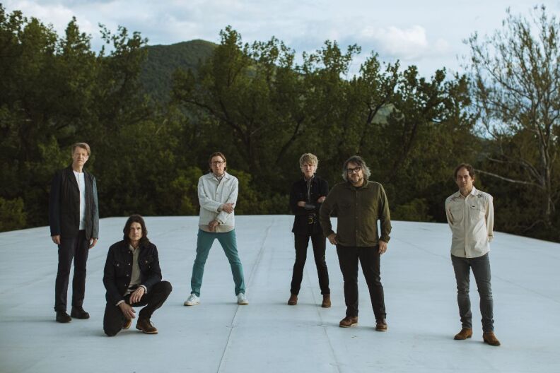 The six members of the band Wilco are pictured  against a backdrop of treetops. One of the members is kneeling while the others stand.