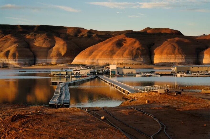 Walkways extend out over lake water. The shore on the near side appears brown and red, and also has some pipes/tubes on it. They extend out to buildings on the water. There are red and brown hills in the background.