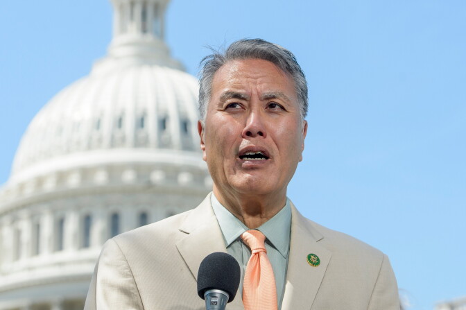 A man with a medium-light skin tone, wearing a white blazer, light blue collared shirt and peach tie, speaks at a microphone with the Capitol building in the background.
