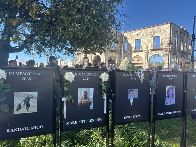 Black posters with photos of Palisades Fire victims are arranged in a row near the shell of a building that burned.