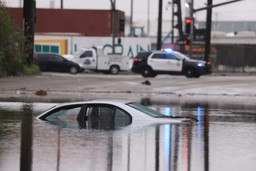 A car sits partially submerged on a flooded road. A police car is visible nearby.