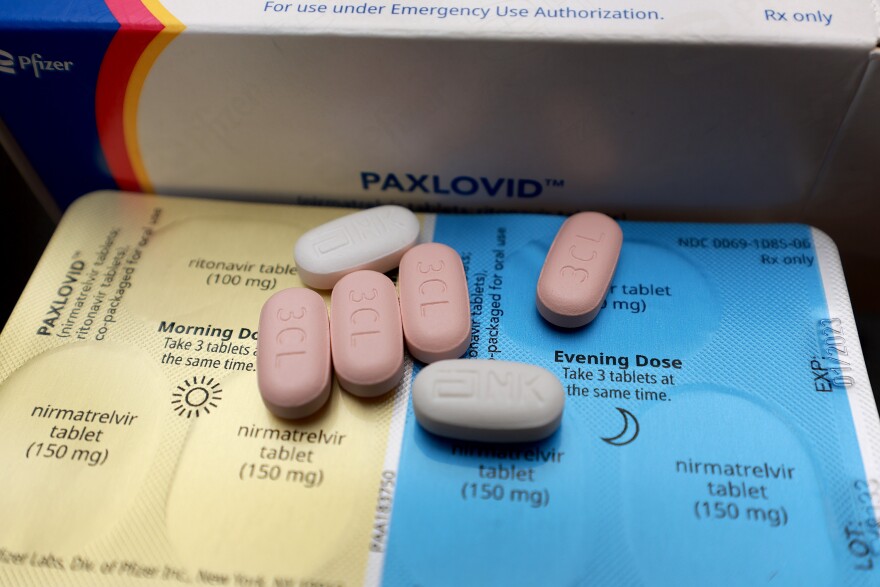 A PAXLOVID box is behind a foil pill holder. There is a yellow side for morning doses and a blue side for evening doses. There are instructions on the container. Six pills lay on the pill holder.