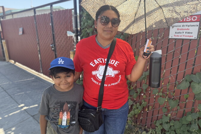 A young boy wearing a blue Dodgers baseball hat stands next to his mother, who is wearing a red shirt and holding an umbrella. Both have medium skin tone. They are standing in front of a chain link fence.