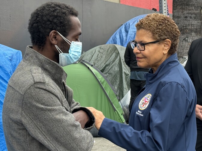 A woman with short hair (right) wearing a blue jacket with the L.A. city seal on it reaches out and holds the arm of a man (left) who's closing his eyes and wearing a mask, in front of tents in the background.