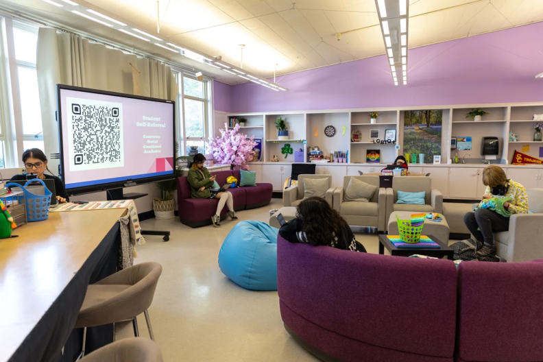 Students are sitting on couches and a table, either doing an activity or on their phones and laptops, in a room with shelves a large monitor with a QR code and text that reads "Student self-referral."