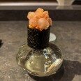 Close-up of a single piece of sushi presented on a decorative glass stand at a restaurant counter. The sushi features pink raw fish or seafood topped with what appears to be uni (sea urchin) or roe, wrapped in dark nori seaweed.