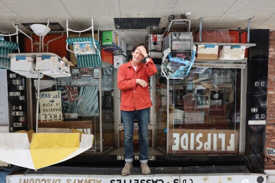 A movie poster for the documentary film "Flipside." It features a man standing in from of a record store that's upside down behind him. He's pressing his hand to his forehead, as if overwhelmed. 