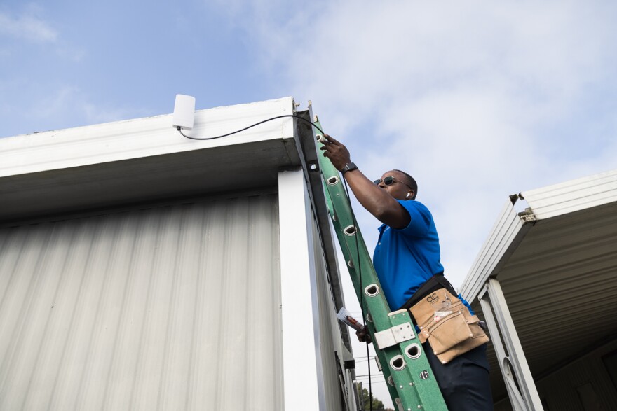 A man with dark brown skin stands on a green ladder as he installs a fixed wireless device on the roof a mobile home.