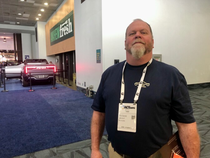A white man with a beard and blue shirt smiles at the camera in a well-lit convention center.  