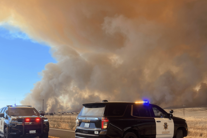 Two black and white law enforcement vehicles are on a road with dark plumes of smoke behind them. 