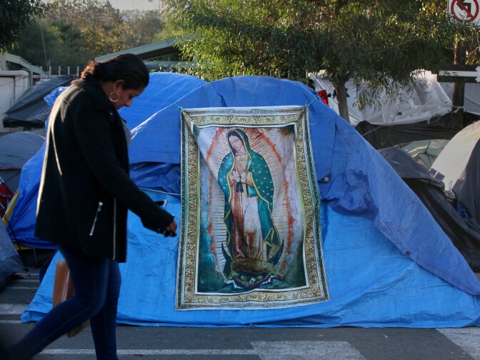 Tijuana, BAJA CALIFORNIA, Mexico - December 16, 2018. At the Benito Juarez Sports Complex near downtown Tijuana,  a woman walks past a tapestry of the Virgen de Guadalupe in the middle of the migrant tents.

 In Tijuana, Mexico, children members of the migrant caravan are learning to live in limbo as they move between shelters, settling in as much as possible to create a sense of normalcy, with help from NGOs, counselors and aid organizations. (Photo by Peggy Peattie)