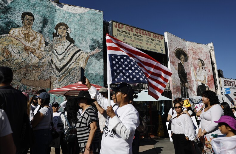 People marching in the street, one man holds up an American flag that is turned upside down. In the background are murals depicting mariachis — and an ice cream shop.