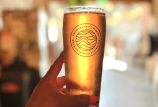 A golden glass of beer has light coming through it. It's being held by a hand. On the glass it says Brewjeria Company, brewed in Pico Rivera