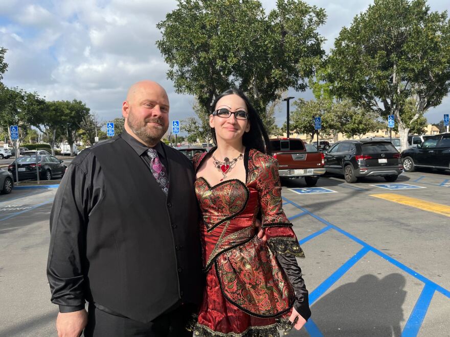 A man with a shaved head, close beard and light-tone skin wears a black shirt and vest with a tie that has hearts on it. His arm is around a woman with light-tone skin in a brocade red dress with a heart-shaped bodice and necklace. She wears glasses and has long straight dark hair.
