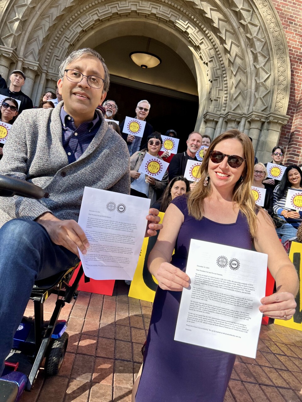 A man, left, and a woman, both with light skin tone, hold pieces of paper. Behind them are a large group of people holding signs with union insignia.
