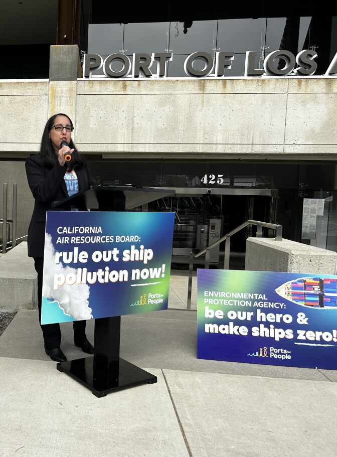 A woman wearing a black suit with dark hair and light brown skin speaks at a podium with a sign reading "California Air Resources Board rule out ship pollution now!"