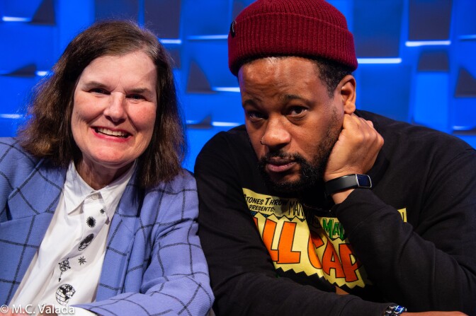 Paula Poundstone, a white woman with a blue blazer and Open Mike Eagle, a dark skinned man with a red stocking cap, looking at the camera
