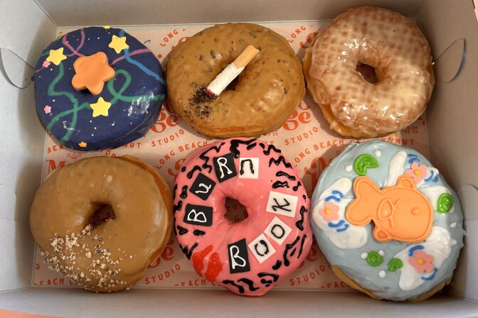 Six doughnuts are lined up in a paper box. The one on the top left has dark blue icing and with yellow stars, the one in the middle has a coffee-brown icing and edible "cigarette" made out of an Italian breadstick. On the bottom, the far right donut is covered in light blue icing and an orange edible fish, with the one next to it covered in pink and black with the words "Burn Book" affixed to the front.