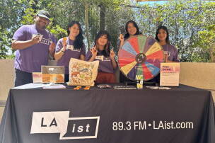 A group of five people hold small items and stand behind a table with a tablecloth that reads "LAist. 89.3 FM. LAist.com" with a spinning wheel and other items on top of it. 