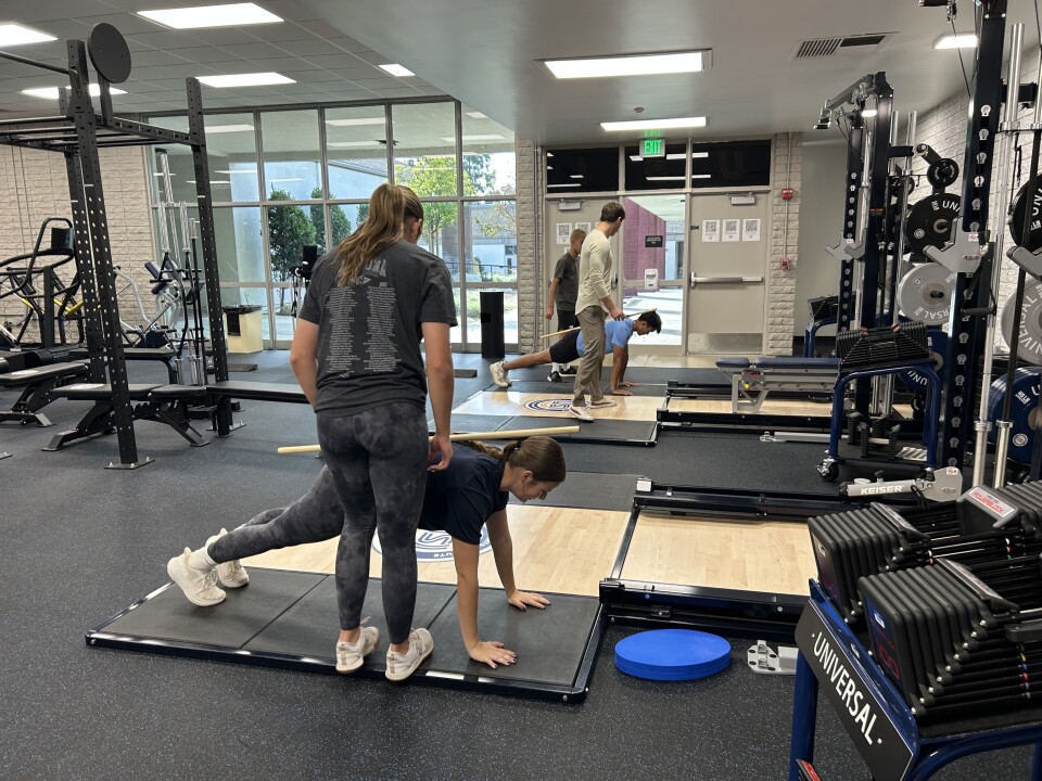 Trainers stand next to several students holding a plank position while balancing wooden poles on their backs. The room is full of gym machines and weights and other physical training equipment.