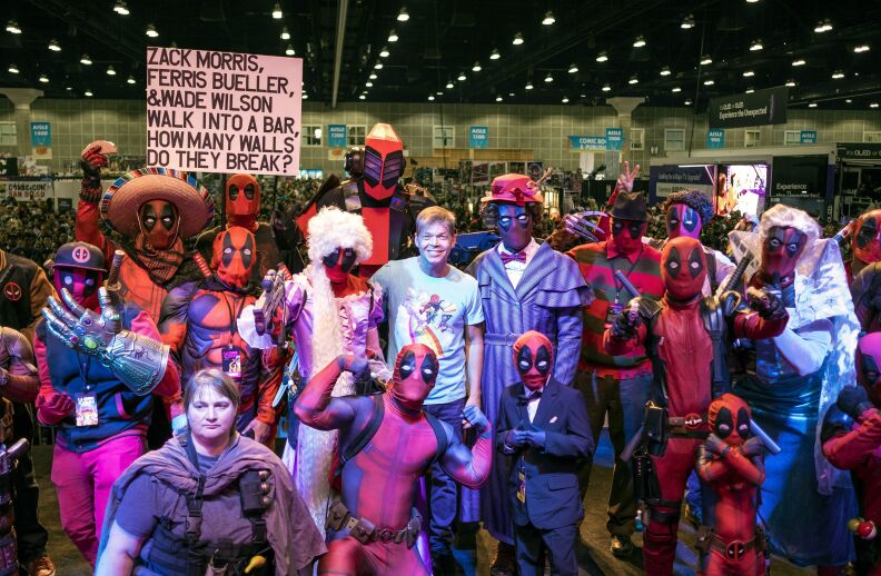 A small crowd of fans wearing various styles of Deadpool costume stand around Deadpool comic co-creator Rob Liefeld. One fan holds up a sign reading "Zack Morris, Ferris Bueller, & Wade Wilson walk into a bar, how many walls do they break?"