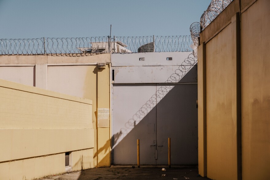 Circles of barbed wire ring the top of tall cement and steel walls painted tepid gray and yellow. A hazy sky is in the background.
