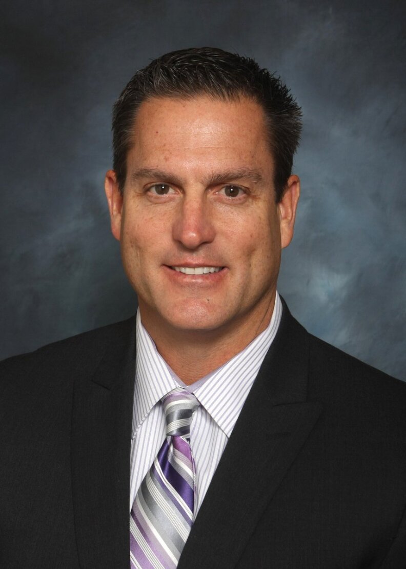 A man with light-tone skin with a square jaw and dimples has short brown hiar and wears a suit with a pin-striped shirt and diagonal striped tie.