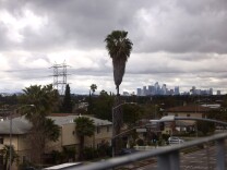 The skyline of Los Angeles in the background surrounded by heavy gray clouds. In the foreground a neighborhood as seen from above. 