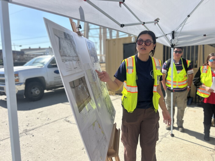 A person in a yellow safety vest points to charts displayed under a tent. Others nearby wear similar vests.