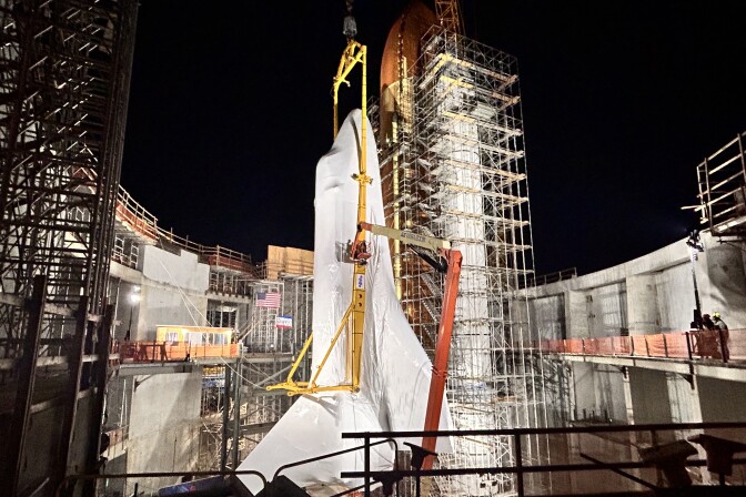 A space shuttle structure towers over the partially-constructed concrete walls and scaffolding that surround it. The structure is displayed vertically towards the pitch black night sky. 