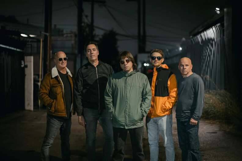 Five men stand in the middle of a dark road. The men pictured in the far left, middle and right of middle all wear sunglasses.