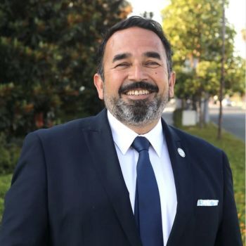 A man with dark hair and a salt-and-pepper beard stands in front of trees wearing a blue suit, white shirt and a blue tie.
